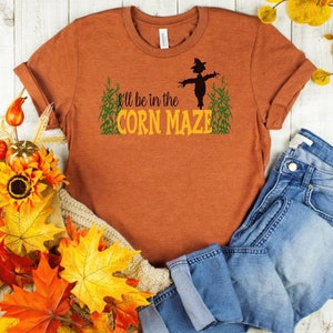 May include: An orange t-shirt with the text "I'll be in the Corn Maze" in green and yellow. A black scarecrow graphic is centered above the text. The shirt is paired with blue jeans and autumn-themed decorations.