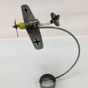 Op de afbeelding: Een metalen sculptuur van een jachtvliegtuig uit de Tweede Wereldoorlog met een gele propeller en zwarte kruisen op de vleugels. Het vliegtuig is gemonteerd op een metalen standaard.