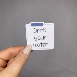 May include: A white sticker with the text "Drink your water" in black, resembling a sticky note. A blue strip is at the top, mimicking tape. The sticker is held against a gray background.