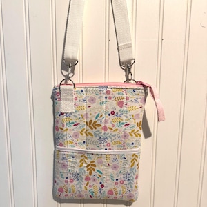 May include: A rectangular crossbody bag featuring a white fabric base with a colorful floral pattern in shades of pink, yellow, and blue. The bag includes a light pink zipper closure, a white exterior pocket, and a white adjustable fabric strap.