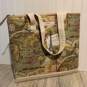 May include: A tote bag featuring a vintage-style world map print in shades of beige, green, and red. The bag has cream-colored fabric handles, a zipper closure, and a solid cream base. Text includes "Around the World" and "Book Your Holidays."