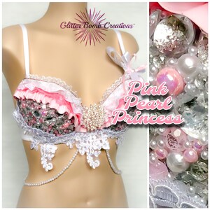 May include: A pink and white bra with lace and pearl embellishments. The bra has a pink ruffle trim and a large rhinestone brooch. The text "Pink Pearl Princess" is visible in the image.