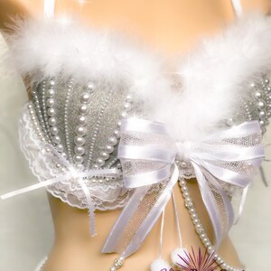 Pu&ograve; includere: Un reggiseno bianco con un ricamo di glitter argento e perle. Il reggiseno ha un fiocco bianco e un bordo di piume bianche. Il reggiseno &egrave; abbinato a una collana di perle bianche e a un girocollo di perle bianche.