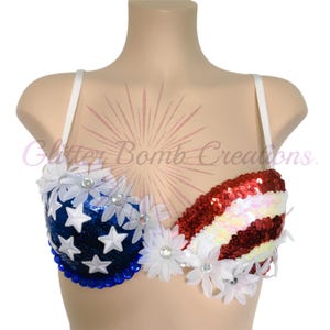May include: A bra featuring a patriotic design with a blue sequined cup adorned with white stars and a red and white striped sequined cup. The garment is decorated with white fabric daisies and blue beads along the bottom edge.