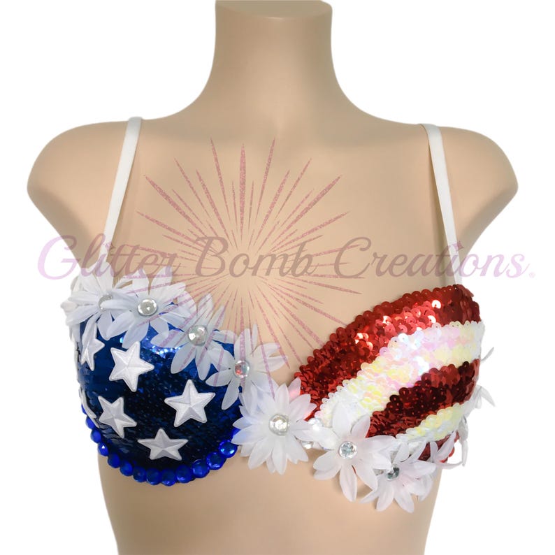 May include: A bra featuring a patriotic design with a blue sequined cup adorned with white stars and a red and white striped sequined cup. The garment is decorated with white fabric daisies and blue beads along the bottom edge.
