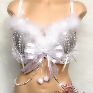 Pu&ograve; includere: Un reggiseno bianco con paillettes argentate e decorazioni di perle, un fiocco bianco e un bordo bianco e soffice. Una catena di perle bianche con pon pon bianchi pende dal reggiseno. Il reggiseno &egrave; su un manichino bianco. Il testo "Glitter Bomb Creations" &egrave; visibile nell'angolo in basso a destra dell'immagine.