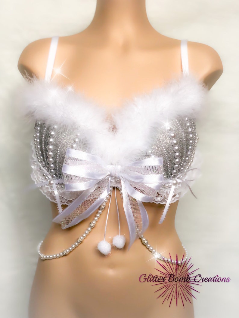 Pu&ograve; includere: Un reggiseno bianco con paillettes argentate e decorazioni di perle, un fiocco bianco e un bordo bianco e soffice. Una catena di perle bianche con pon pon bianchi pende dal reggiseno. Il reggiseno &egrave; su un manichino bianco. Il testo "Glitter Bomb Creations" &egrave; visibile nell'angolo in basso a destra dell'immagine.
