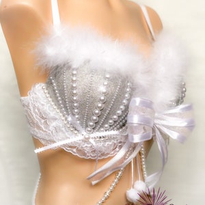 Pu&ograve; includere: Un reggiseno bianco con decorazioni in glitter argento e perle. Il reggiseno ha un bordo in pelliccia bianca e un fiocco di raso bianco. Una collana di perle con pon pon bianchi pende dal reggiseno. Il testo "Glitter Bomb Creations" &egrave; visibile nell'angolo in basso a destra dell'immagine.