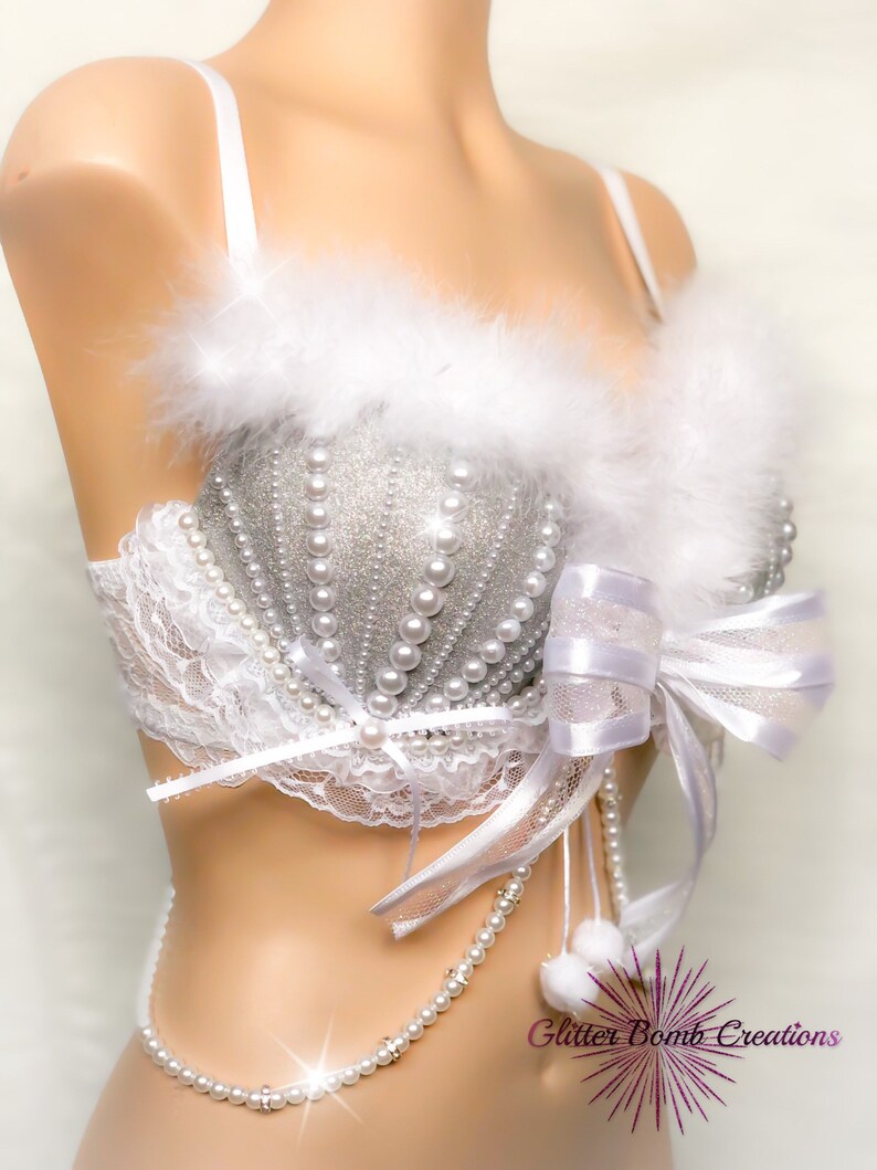 Pu&ograve; includere: Un reggiseno bianco con decorazioni in glitter argento e perle. Il reggiseno ha un bordo in pelliccia bianca e un fiocco di raso bianco. Una collana di perle con pon pon bianchi pende dal reggiseno. Il testo "Glitter Bomb Creations" &egrave; visibile nell'angolo in basso a destra dell'immagine.