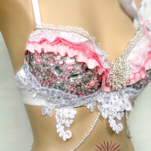 May include: A white and pink bra with lace trim and pearl embellishments. The bra has a pink ribbon and a white bow on the strap. The bra is on a white mannequin.
