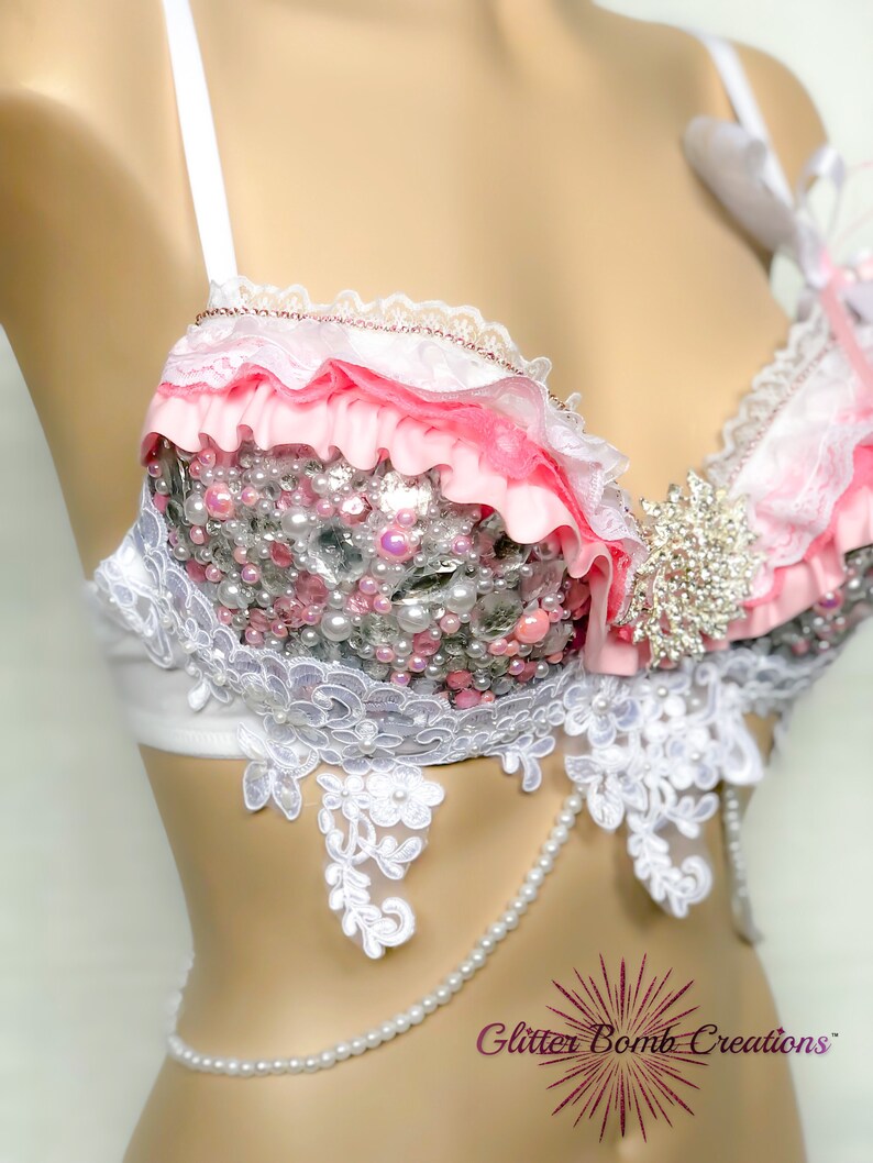 May include: A white and pink bra with lace trim and pearl embellishments. The bra has a pink ribbon and a white bow on the strap. The bra is on a white mannequin.