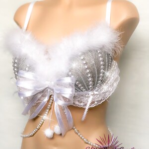 Pu&ograve; includere: Reggiseno bianco con un design di glitter argento e perle. Il reggiseno ha un bordo in pelliccia bianca e un fiocco bianco con una catena di perle attaccata. Il reggiseno &egrave; su un manichino bianco. Il testo "Glitter Bomb Creations" &egrave; visibile nell'angolo in basso a destra dell'immagine.