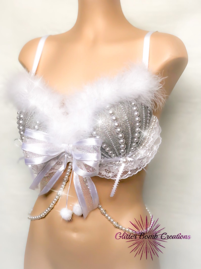 Pu&ograve; includere: Reggiseno bianco con un design di glitter argento e perle. Il reggiseno ha un bordo in pelliccia bianca e un fiocco bianco con una catena di perle attaccata. Il reggiseno &egrave; su un manichino bianco. Il testo "Glitter Bomb Creations" &egrave; visibile nell'angolo in basso a destra dell'immagine.