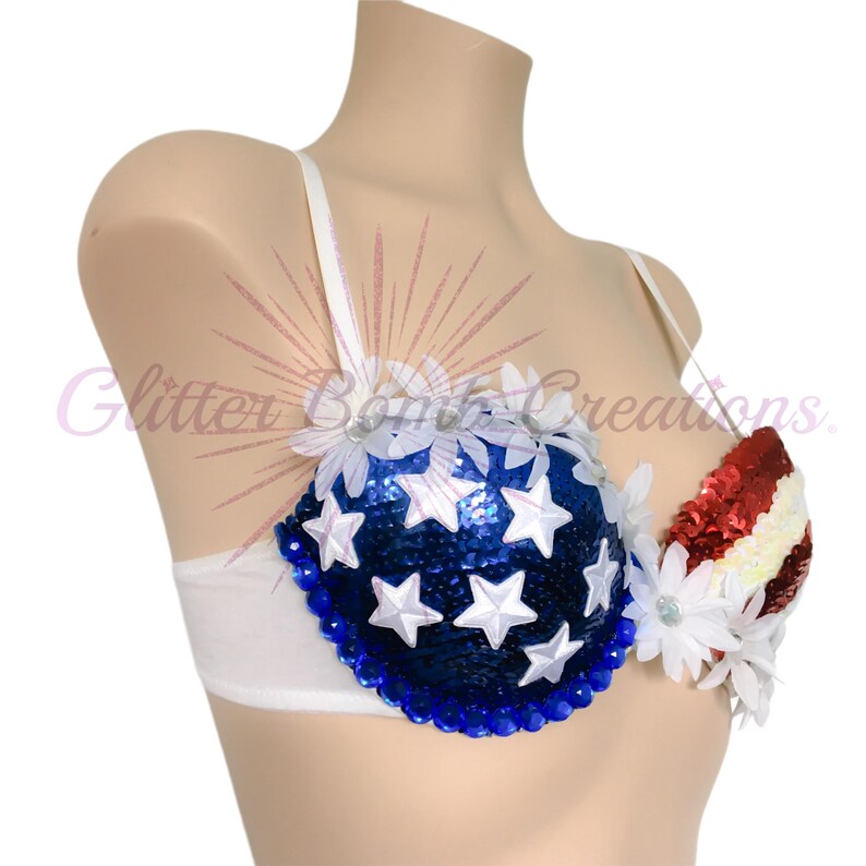 May include: A white bra featuring a blue sequined cup adorned with white stars and white floral accents, and a red, white, and blue striped sequined cup. The text Glitter Bomb Creations is overlaid on the image in a light purple script font.