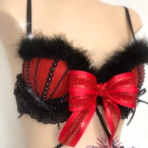 May include: Red glitter bra with black feather trim, black pearl accents, and a large red bow. The bra features a beaded strap and is designed with a decorative, eye-catching style.