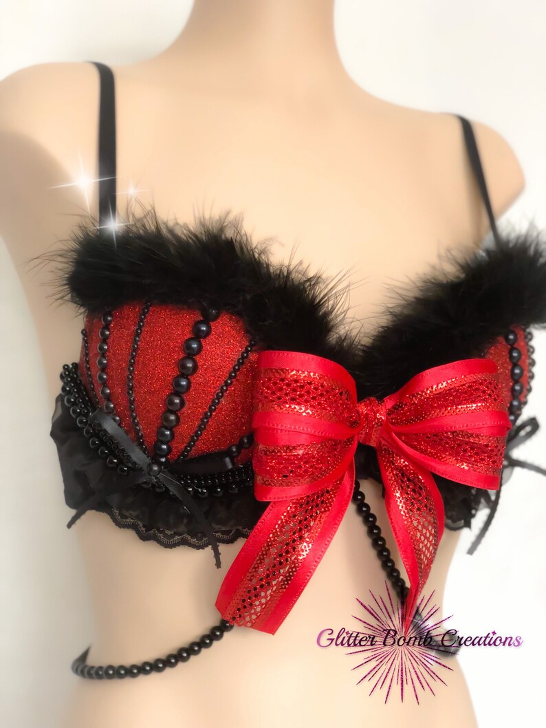 May include: Red glitter bra with black feather trim, black pearl accents, and a large red bow. The bra features a beaded strap and is designed with a decorative, eye-catching style.