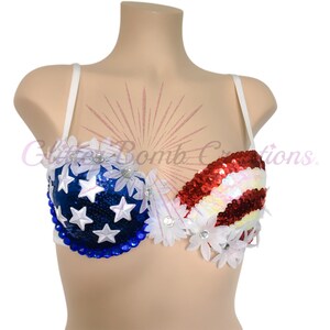 May include: A patriotic bra featuring a blue sequined cup with white stars and a red and white striped sequined cup. The garment is decorated with white fabric flowers and blue beads along the bottom edge, with white straps on a mannequin.