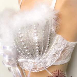 Pu&ograve; includere: Un reggiseno bianco con un ricamo di glitter argento e perle. Il reggiseno ha un bordo in pizzo bianco e un bordo in piume bianche. Il reggiseno &egrave; su un manichino bianco.