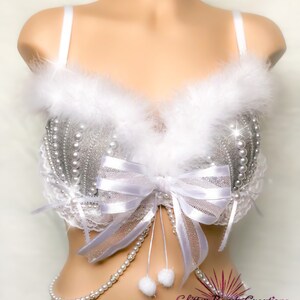 Pu&ograve; includere: Un reggiseno bianco con decorazioni in glitter argento e perle, un fiocco bianco e un bordo in piume bianche. Il reggiseno ha una catena di perle bianche attaccata alla parte inferiore. Il testo "Glitter Bomb Creations" &egrave; visibile nell'angolo in basso a destra dell'immagine.