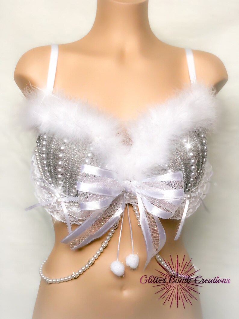 Pu&ograve; includere: Un reggiseno bianco con decorazioni in glitter argento e perle, un fiocco bianco e un bordo in piume bianche. Il reggiseno ha una catena di perle bianche attaccata alla parte inferiore. Il testo "Glitter Bomb Creations" &egrave; visibile nell'angolo in basso a destra dell'immagine.