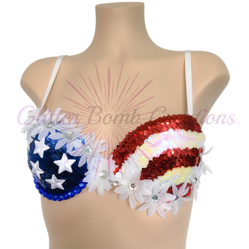 May include: A sequined bra featuring a blue cup with white stars and a red and white striped cup. The garment is decorated with white daisy-shaped floral appliqu&eacute;s and clear rhinestones. The text Glitter Bomb Creations is visible in the center.