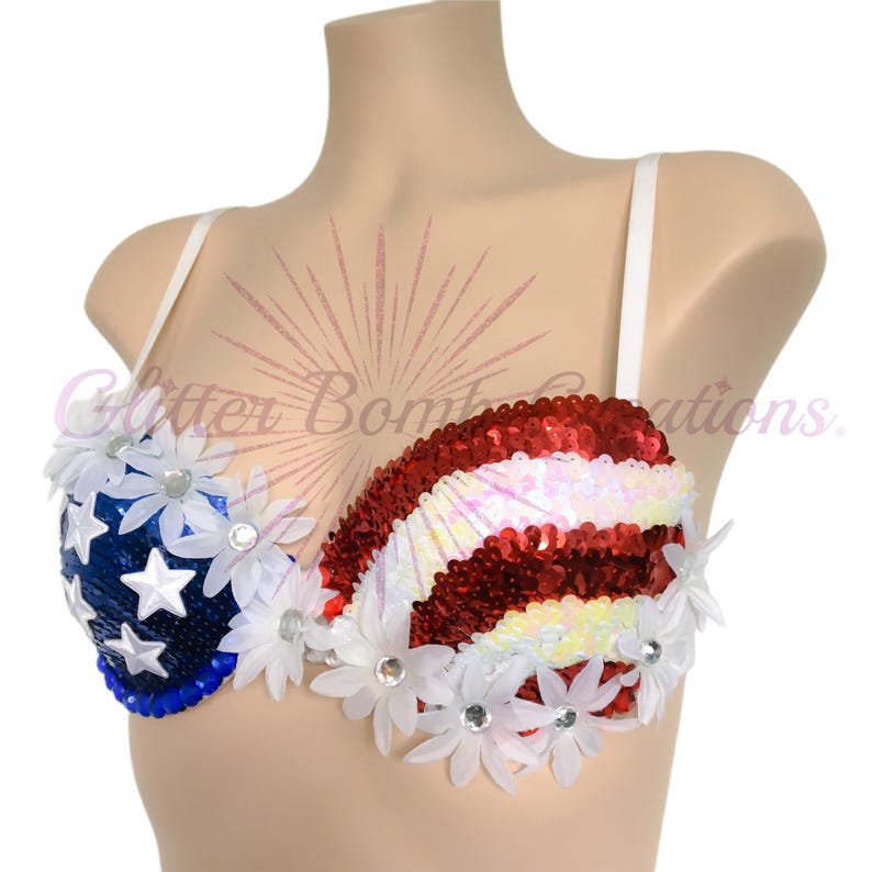May include: A sequined bra featuring a patriotic design with a blue section adorned with white stars and a red and white striped section. The garment is embellished with white daisy-shaped floral appliqu&eacute;s with clear centers and white straps.