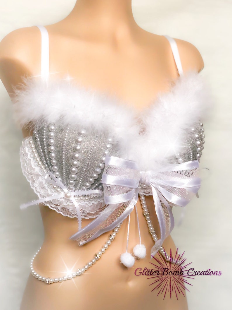 Pu&ograve; includere: Un reggiseno bianco e argento con un bordo di pelliccia bianca, un fiocco bianco e una catena di perle. Il reggiseno &egrave; in tessuto glitterato argento e perle bianche.