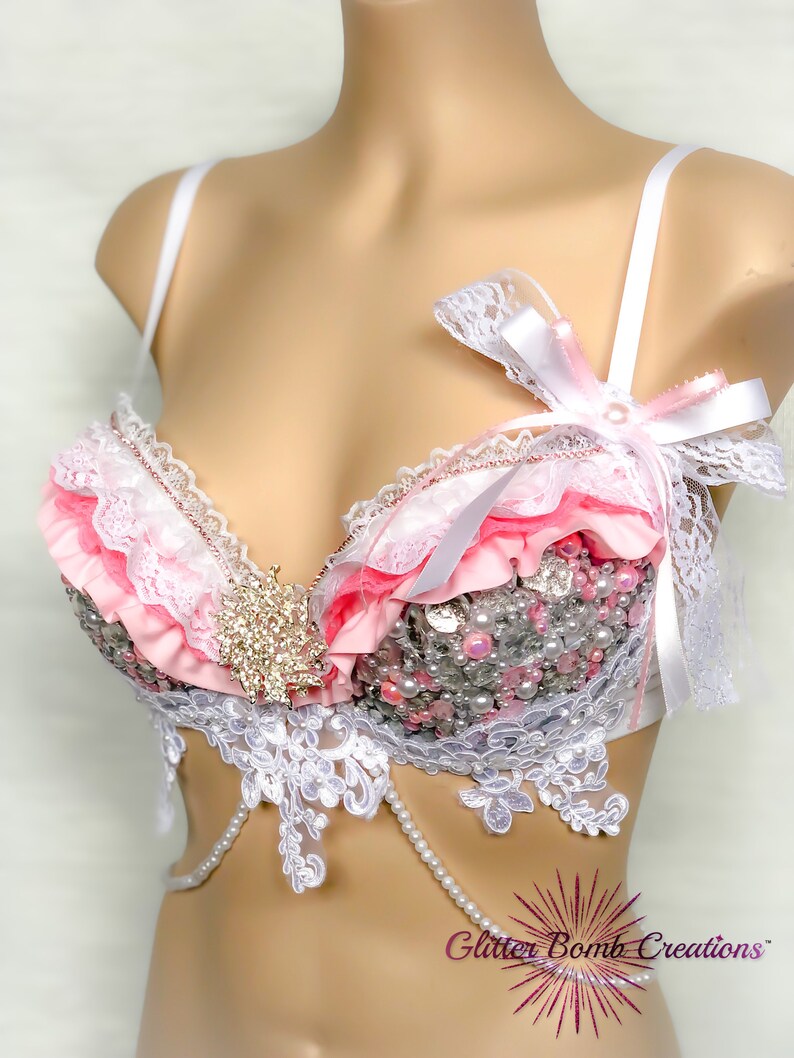 May include: A white and pink lace bra with a pearl and rhinestone embellishment. The bra has a white satin bow on the strap.