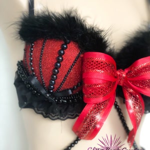 May include: A red glittery bra with black accents. The bra features black feathers, black beaded trim, and a large red bow. The bra has a decorative beaded chain and black lace trim. The bra is a unique, handmade item.