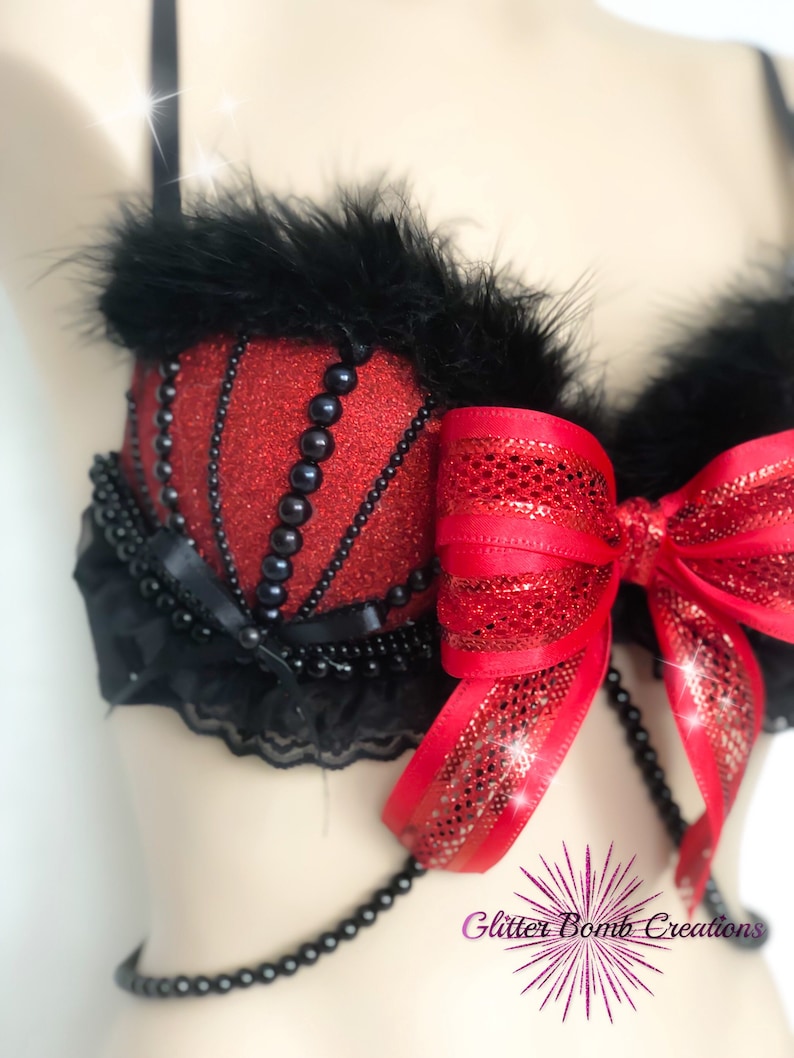 May include: A red glittery bra with black accents. The bra features black feathers, black beaded trim, and a large red bow. The bra has a decorative beaded chain and black lace trim. The bra is a unique, handmade item.