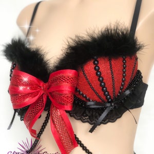 May include: Red and black embellished bra with a bow and bead accents. The bra features a red glitter fabric with black bead detailing and black feather trim along the top edge. A large red bow is centered on the front.