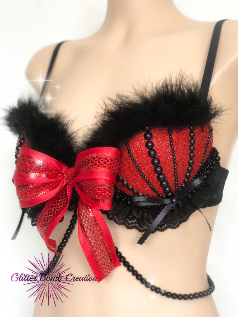 May include: Red and black embellished bra with a bow and bead accents. The bra features a red glitter fabric with black bead detailing and black feather trim along the top edge. A large red bow is centered on the front.