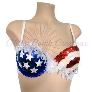 May include: A white bra featuring a patriotic design with blue sequins and white stars on one cup, and red and white sequined stripes on the other. The garment is decorated with white fabric daisies and blue gems along the bottom edge.