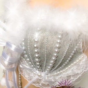Pu&ograve; includere: Un reggiseno bianco e argento con un designo di perle e glitter. Il reggiseno ha un bordo in pizzo bianco e un bordo in piume bianche. Il reggiseno &egrave; su un manichino bianco.