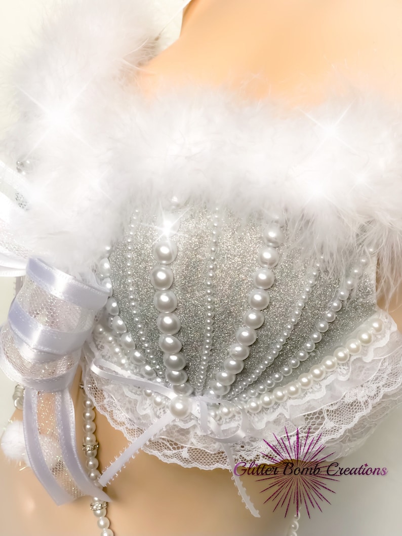 Pu&ograve; includere: Un reggiseno bianco e argento con un designo di perle e glitter. Il reggiseno ha un bordo in pizzo bianco e un bordo in piume bianche. Il reggiseno &egrave; su un manichino bianco.