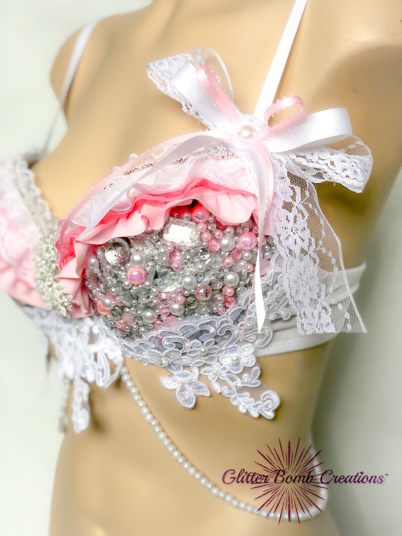 May include: A white and pink lingerie bra with lace, ruffles, and pearl embellishments. The bra has a white ribbon tied into a bow at the top.