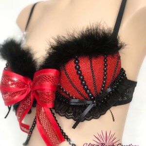May include: A red glitter bra with black feather trim and black bead accents. A large red bow adorns the front, and a beaded strand hangs below. The bra has black straps and lace detailing.
