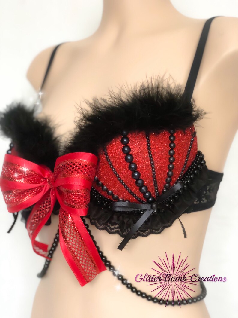May include: A red glitter bra with black feather trim and black bead accents. A large red bow adorns the front, and a beaded strand hangs below. The bra has black straps and lace detailing.