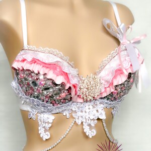 May include: A pink and white lingerie bra with lace and pearl embellishments. The bra has a ruffled trim and a large rhinestone embellishment in the centre. The bra is on a white mannequin.