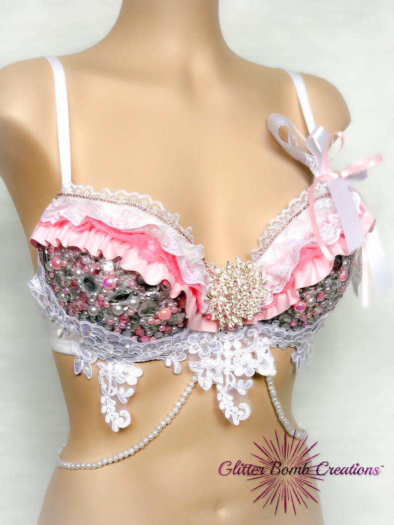 May include: A pink and white lingerie bra with lace and pearl embellishments. The bra has a ruffled trim and a large rhinestone embellishment in the centre. The bra is on a white mannequin.