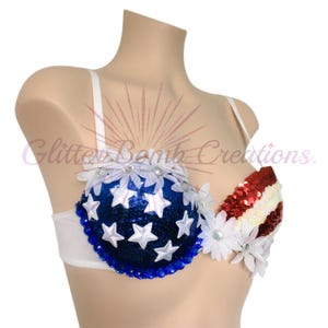 May include: A white bra featuring a blue sequined cup adorned with white stars and blue gems, and a red and white striped sequined cup. The design includes white fabric flowers with center crystals along the top edges of the cups.
