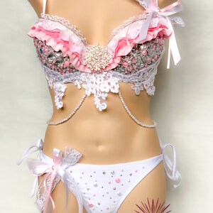 Op de afbeelding: Een witte lingerie set met roze en witte ruches, kant en parelversieringen. De bh heeft een sweetheart-halslijn en verstelbare bandjes. De bijpassende slip heeft een lage taille en een delicate kanten rand. De set is versierd met strass en parels.