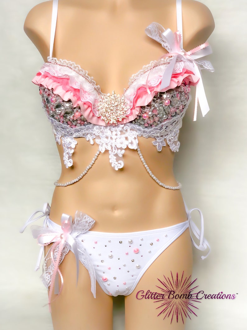 Op de afbeelding: Een witte lingerie set met roze en witte ruches, kant en parelversieringen. De bh heeft een sweetheart-halslijn en verstelbare bandjes. De bijpassende slip heeft een lage taille en een delicate kanten rand. De set is versierd met strass en parels.