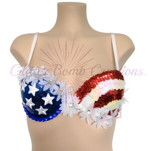 May include: A bra featuring a patriotic design with blue sequins and white stars on one side and red and white sequined stripes on the other. The garment is embellished with white fabric flowers and clear rhinestones. Text reads Glitter Bomb Creations.