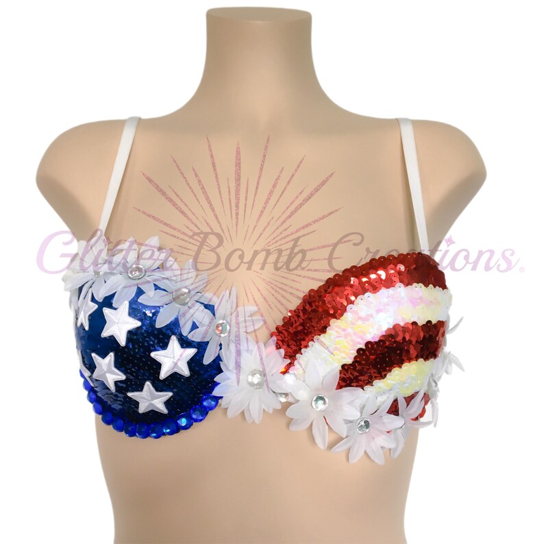 May include: A bra featuring a patriotic design with blue sequins and white stars on one side and red and white sequined stripes on the other. The garment is embellished with white fabric flowers and clear rhinestones. Text reads Glitter Bomb Creations.