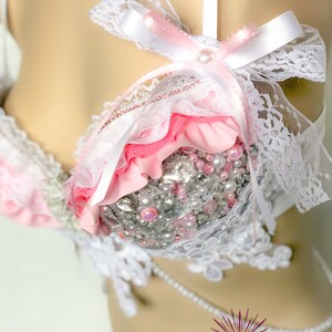 May include: A pink and white lace bra with pearl and rhinestone embellishments. The bra has a white satin bow with a pearl in the centre.