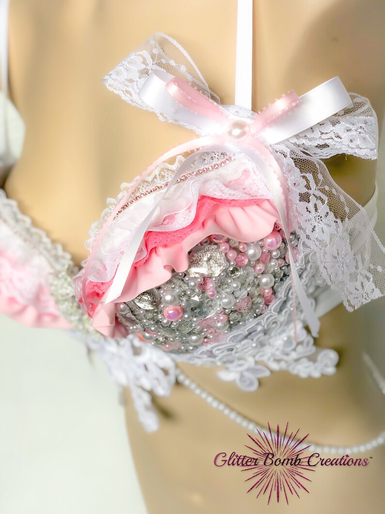 May include: A pink and white lace bra with pearl and rhinestone embellishments. The bra has a white satin bow with a pearl in the centre.