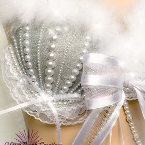 Pu&ograve; includere: Un reggiseno bianco con una coppa in glitter argento, perle bianche e un fiocco di raso bianco. Il reggiseno ha un bordo in pizzo bianco e un bordo in piume bianche.