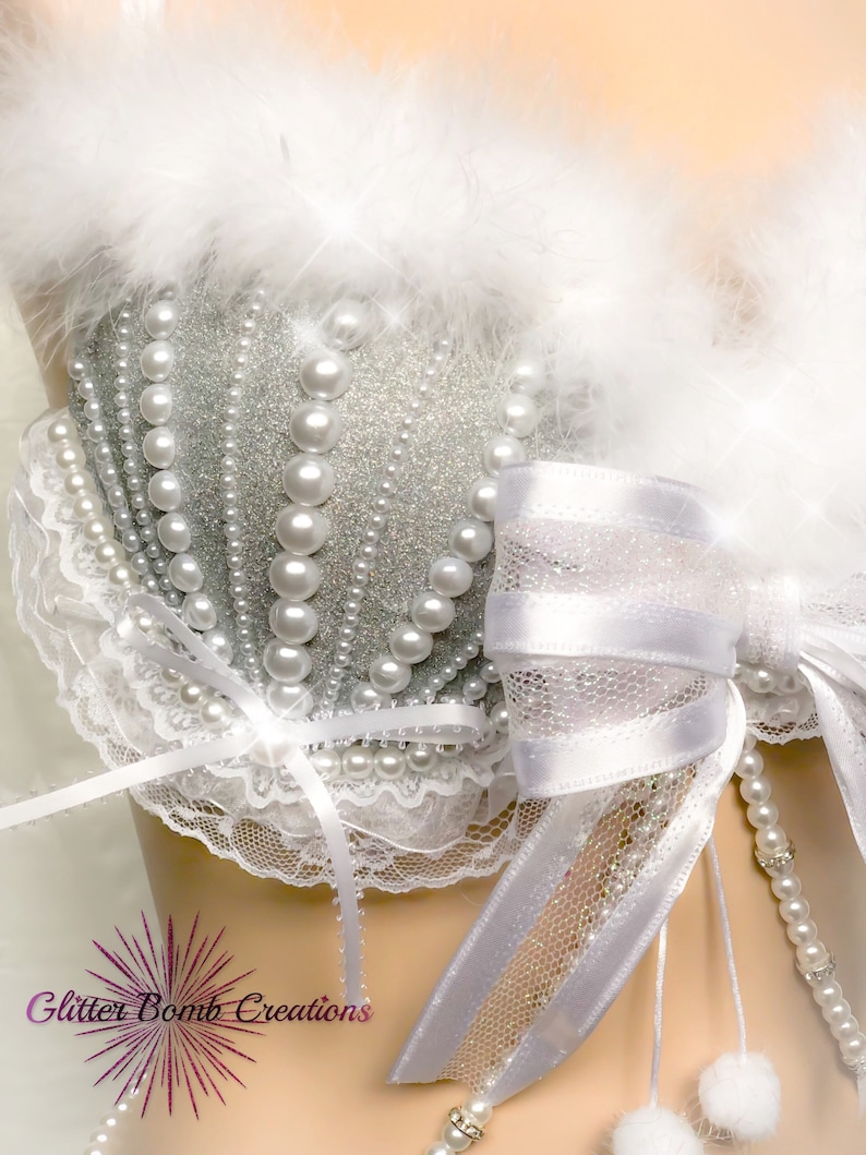 Pu&ograve; includere: Un reggiseno bianco con una coppa in glitter argento, perle bianche e un fiocco di raso bianco. Il reggiseno ha un bordo in pizzo bianco e un bordo in piume bianche.