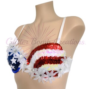 May include: A bra featuring a red, white, and blue sequined pattern resembling the American flag. The left cup is blue with white stars, while the right cup has red and white stripes. White fabric flowers with center crystals are attached.